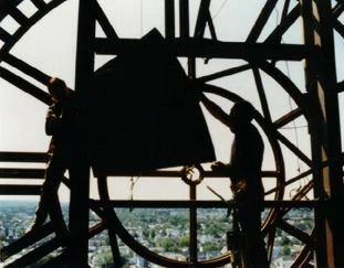 Two workers silhouetted inside a clock tower behind the skeletal dial, the town far below.